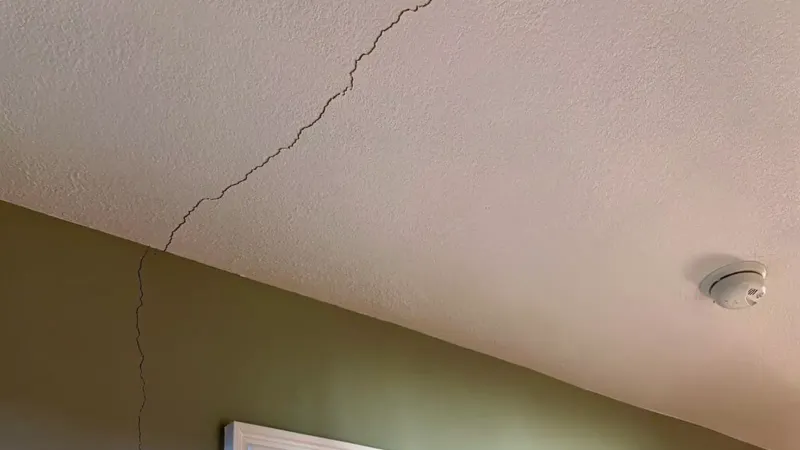 A fine hairline crack running across a lightly textured painted ceiling toward the corner where the ceiling meets the wall, with a round white smoke detector mounted nearby