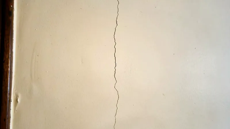 A fine vertical hairline crack running up a cream painted interior wall beside a narrow dark-wood door casing