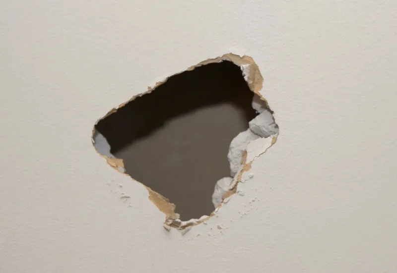 Jagged impact hole broken through a painted interior drywall wall, torn paper facing and crumbling gypsum around the opening exposing the dark cavity behind