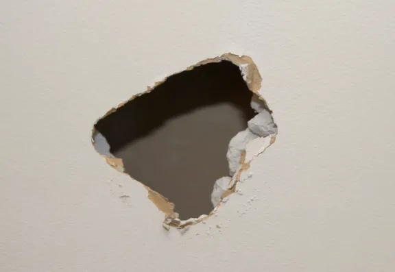 Jagged impact hole broken through a painted interior drywall wall, torn paper facing and crumbling gypsum around the opening exposing the dark cavity behind