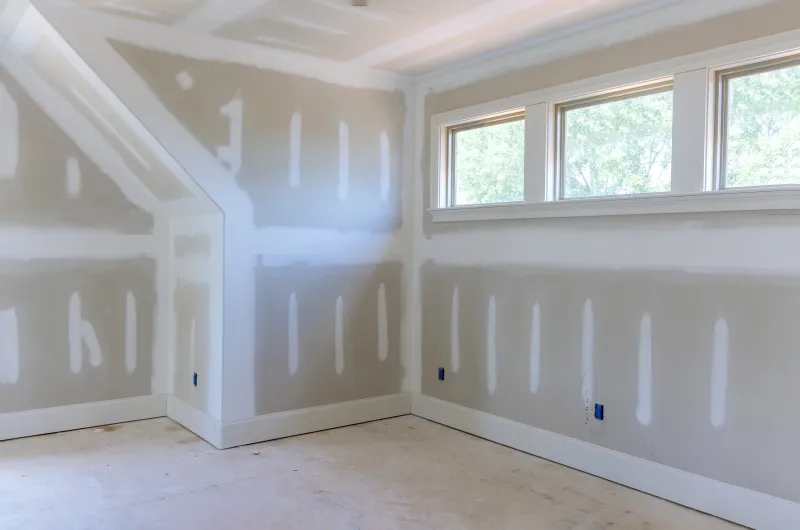 Room with a sloped dormer ceiling in drywall, seams taped and coated and fasteners spotted in white compound over grey board, baseboard and window casing already installed below a bank of three windows