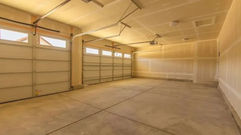 Two-bay garage with the walls and ceiling drywalled and taped, overhead doors closed and a bare concrete floor