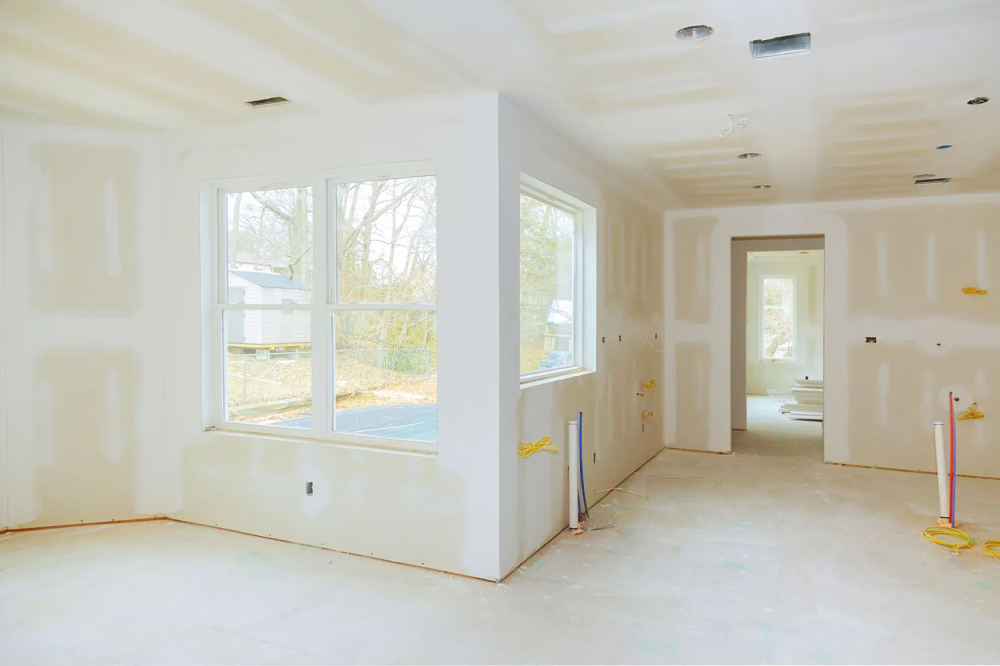 Bright empty room with primed and taped drywall walls and ceiling, a corner bay of windows, and an open doorway to further rooms before trim