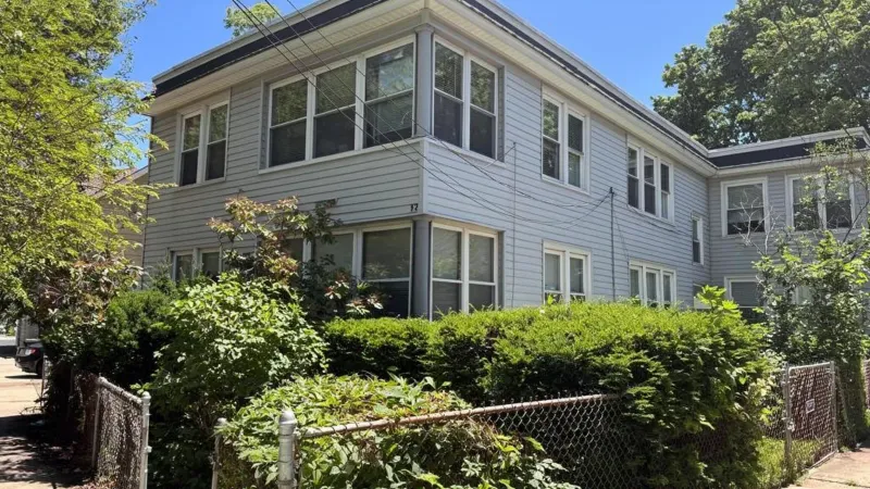 A two-story multi-family house with pale clapboard siding behind a trimmed hedge and chain-link fence