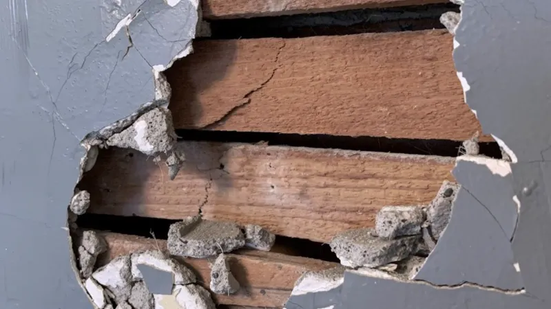 Hole broken through a painted plaster wall, exposing the horizontal wood lath behind it and the crumbling plaster keys around the opening
