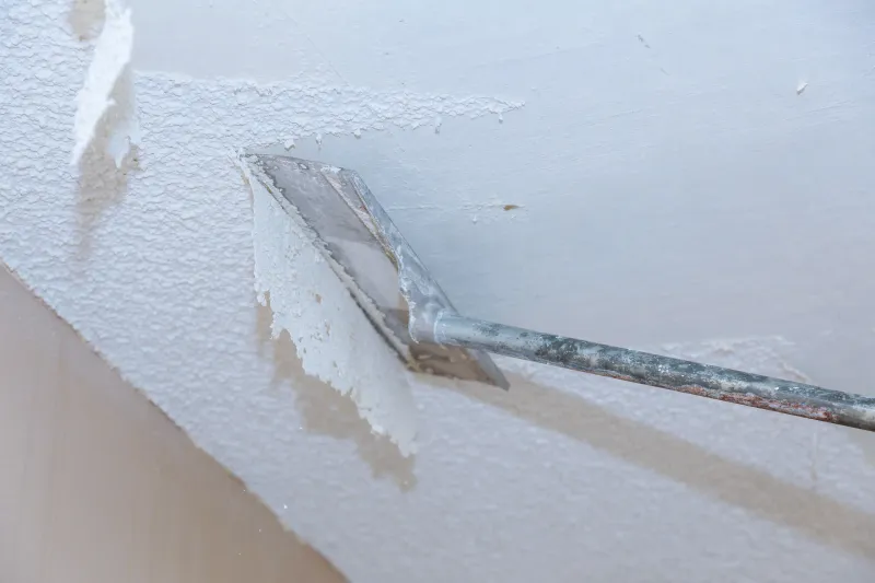 Close-up of a scraper blade lifting popcorn texture off a ceiling, the stripped surface smooth behind it